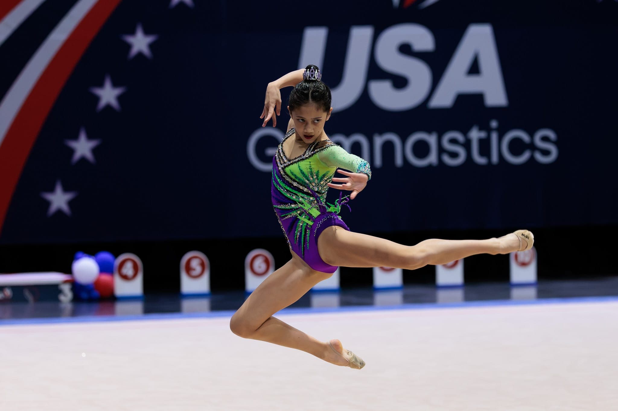 A Boston Rhythmic gymnast in a purple and green leotard mid-leap during a competition routine.