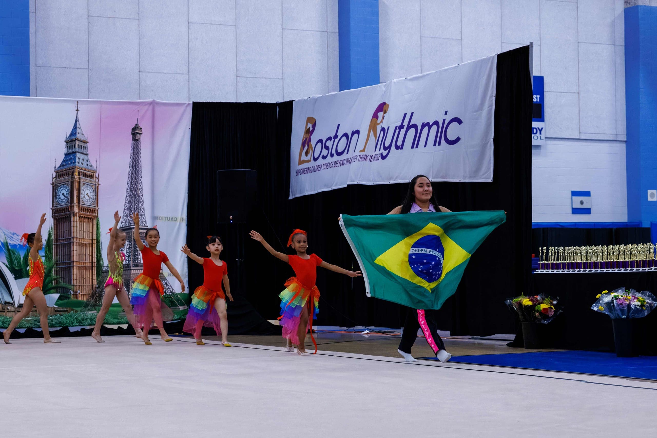 Young Boston Rhythmic gymnasts in rainbow costumes performing while a student carries the Brazil flag at an Around the World show.