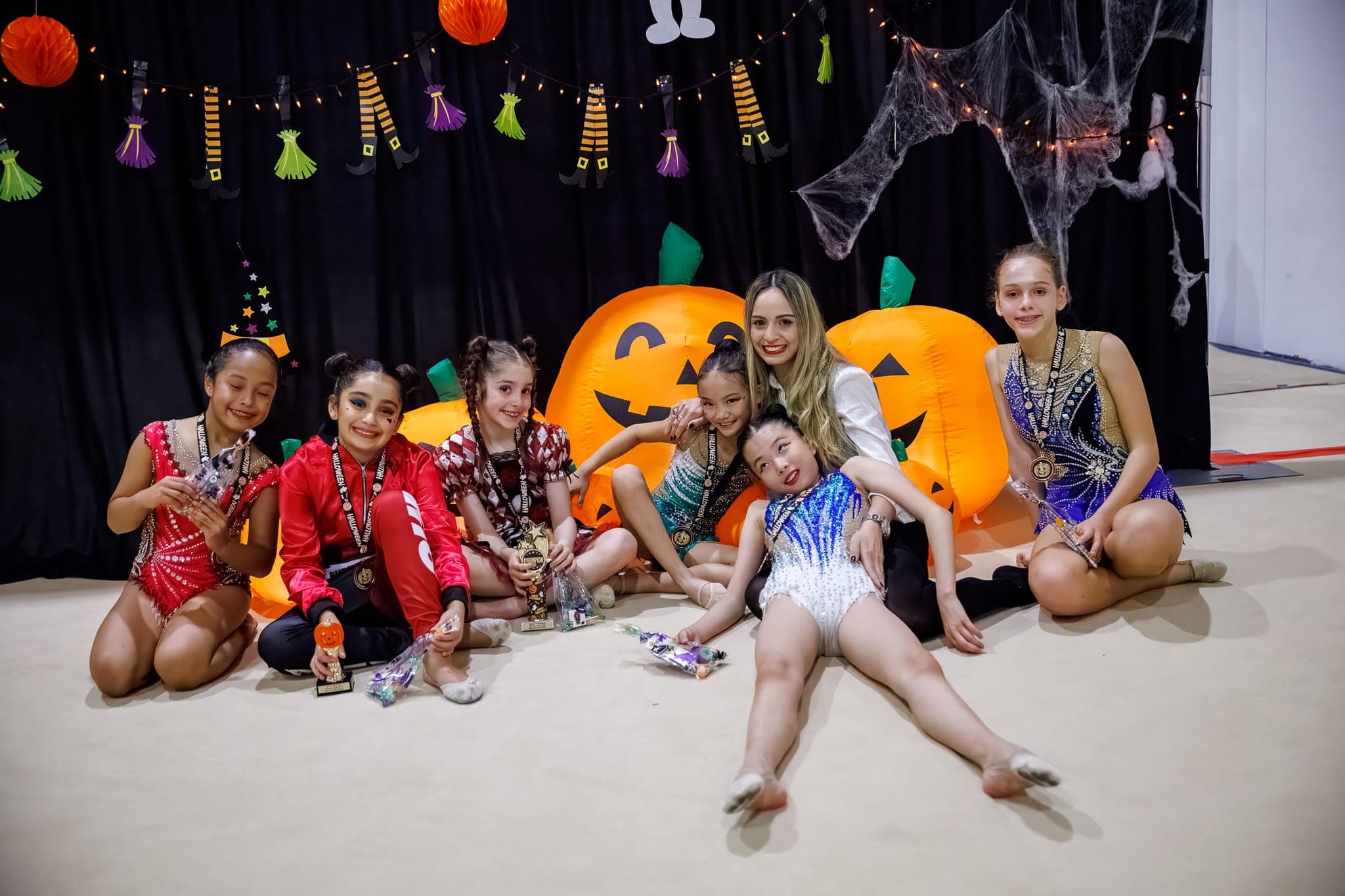 A group of Boston Rhythmic gymnasts and their coach smiling with medals and trophies in front of Halloween pumpkin decorations at the Spooky Cup.