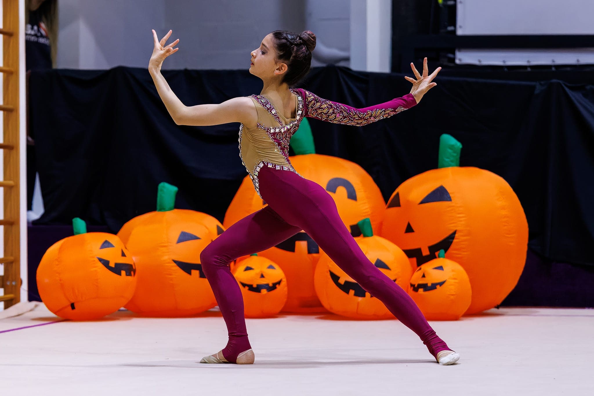 A Boston Rhythmic gymnast in a maroon and gold leotard performing a dramatic pose in front of inflatable jack-o'-lantern pumpkins at the Pumpkin Cup.