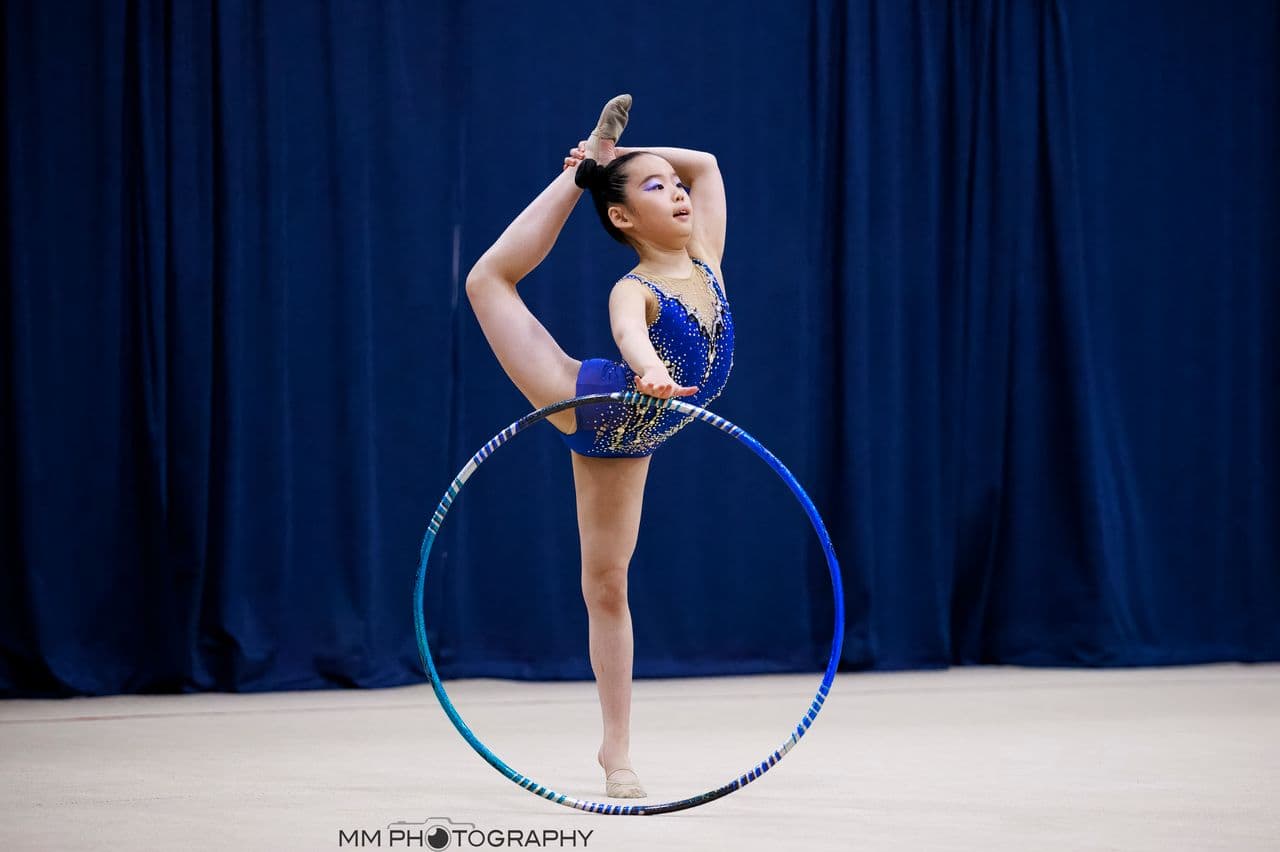 A Boston Rhythmic gymnast in a blue leotard holding a standing split while balancing a hoop on the floor at the Friendship Cup.