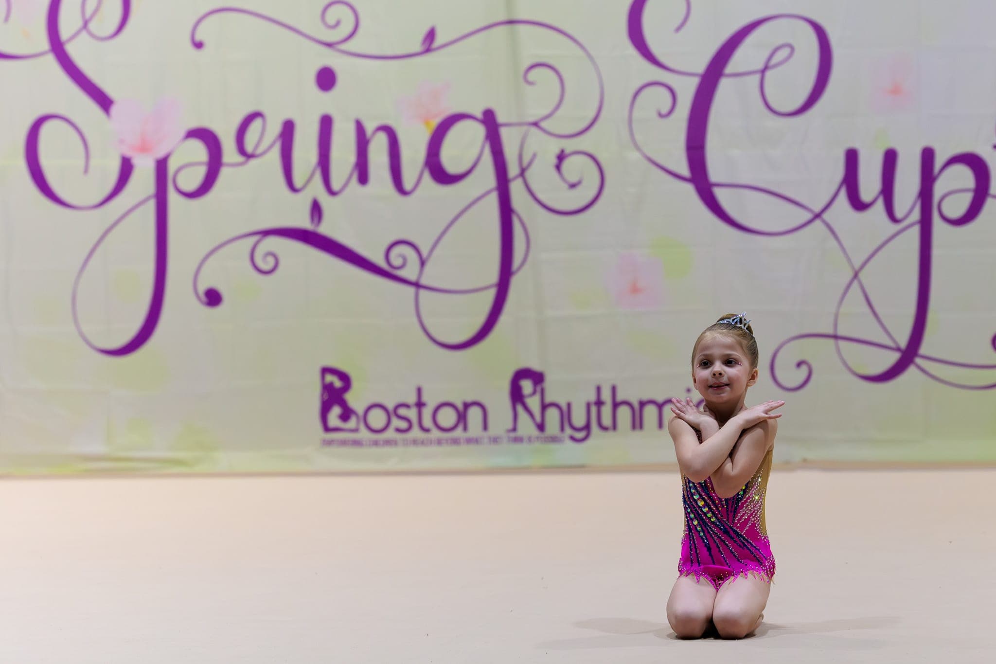A young Boston Rhythmic gymnast in a sparkly pink and gold leotard and tiara kneeling on the floor in front of the Spring Cup banner.