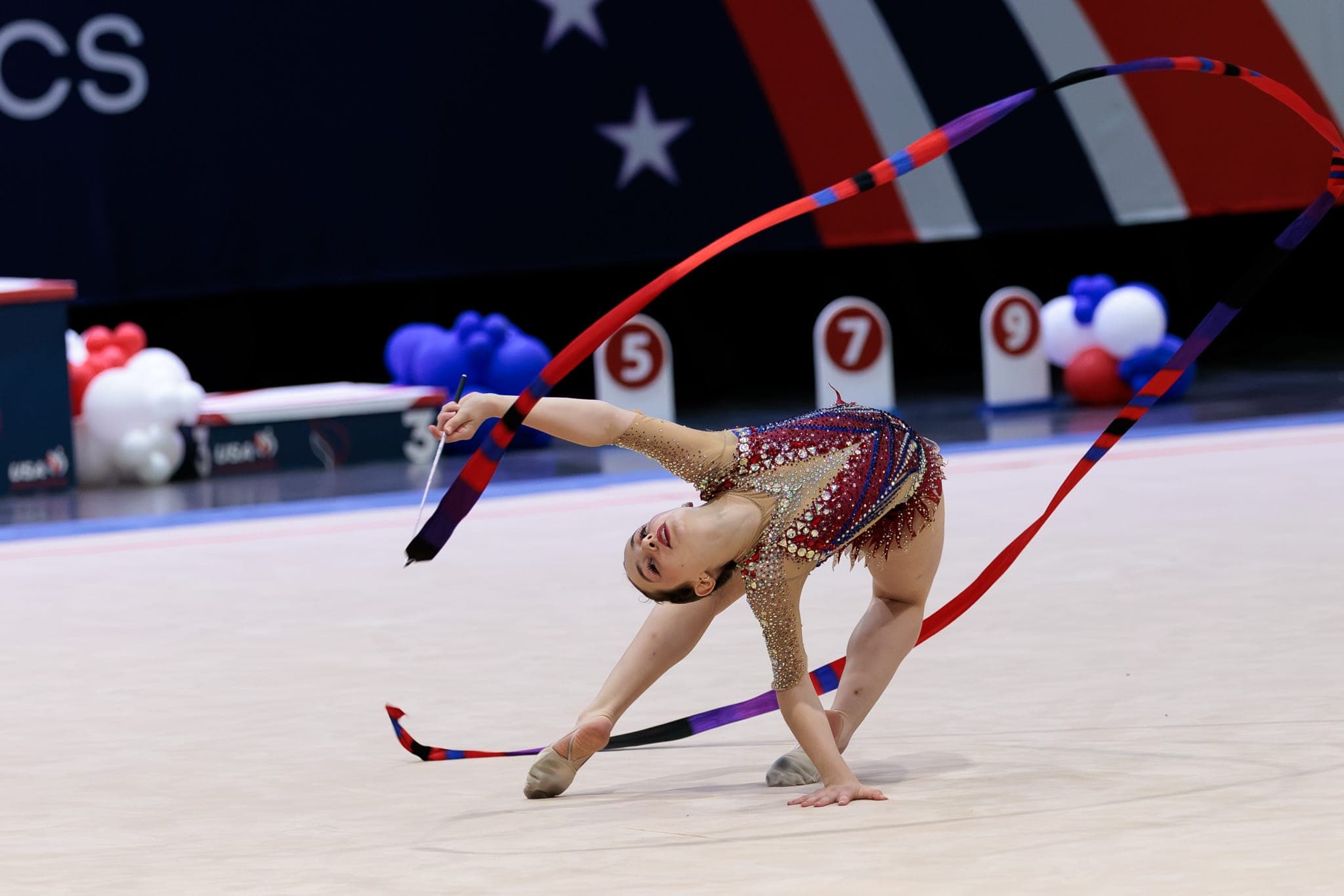 A Boston Rhythmic gymnast in a deep backbend trailing a red-and-blue ribbon on the competition floor.