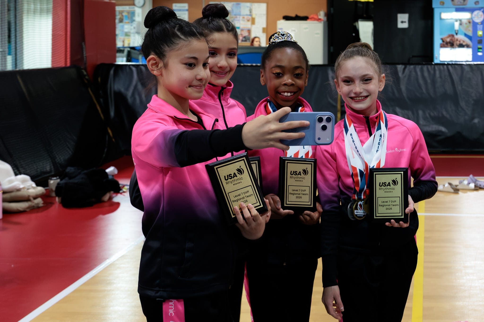 Four young Boston Rhythmic gymnasts in pink team jackets taking a selfie while holding regional team plaques.