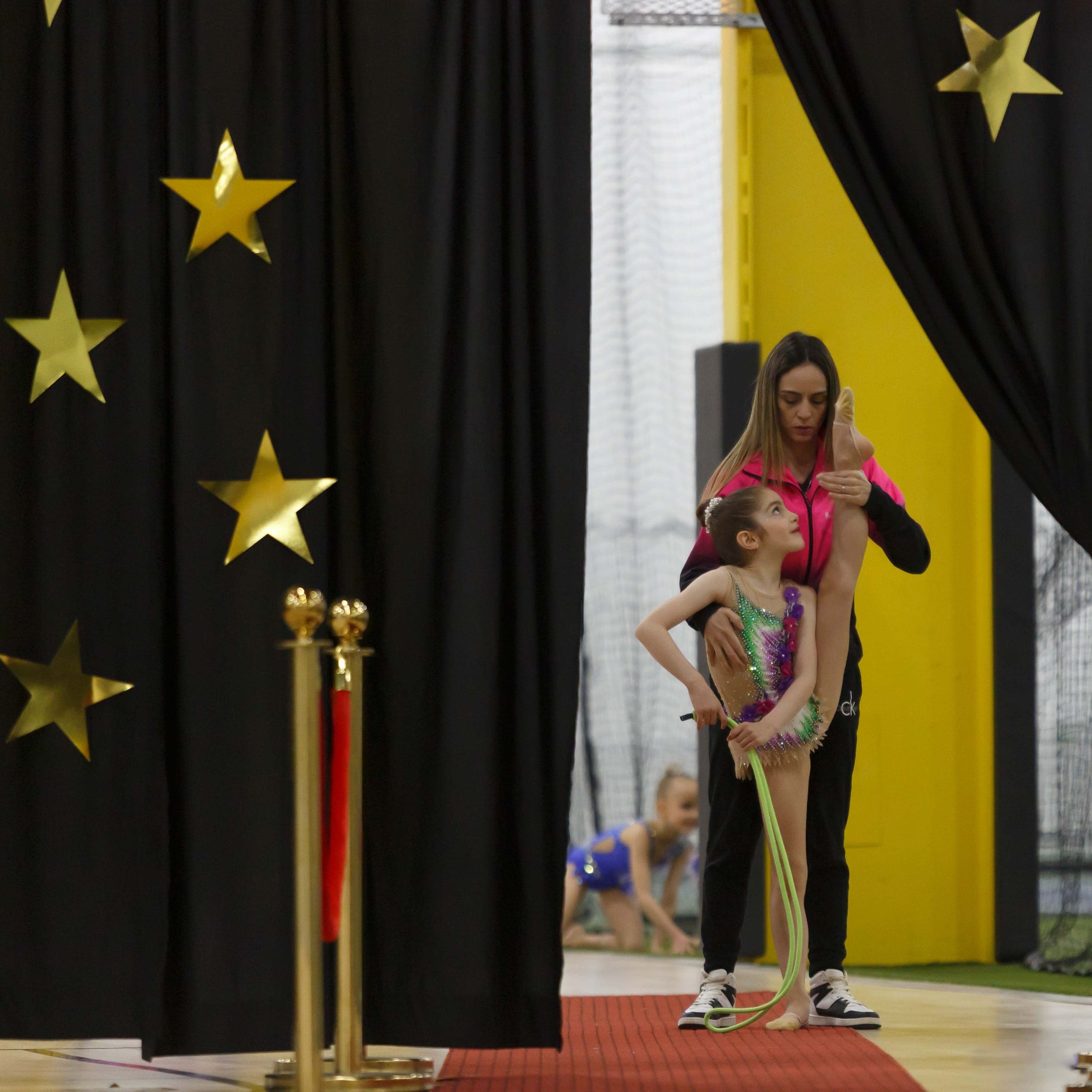 A Boston Rhythmic coach in a pink jacket supporting a young gymnast with a jump rope backstage behind star-decorated curtains.