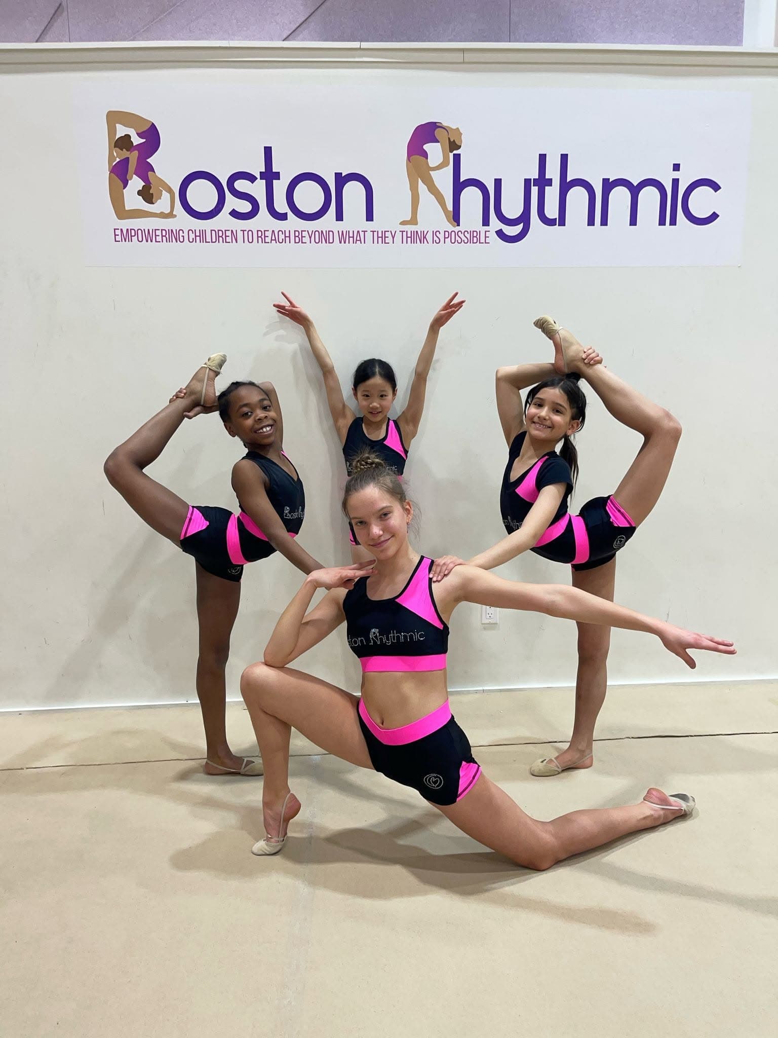 Boston Rhythmic gymnasts posing in flexibility positions in front of the studio logo